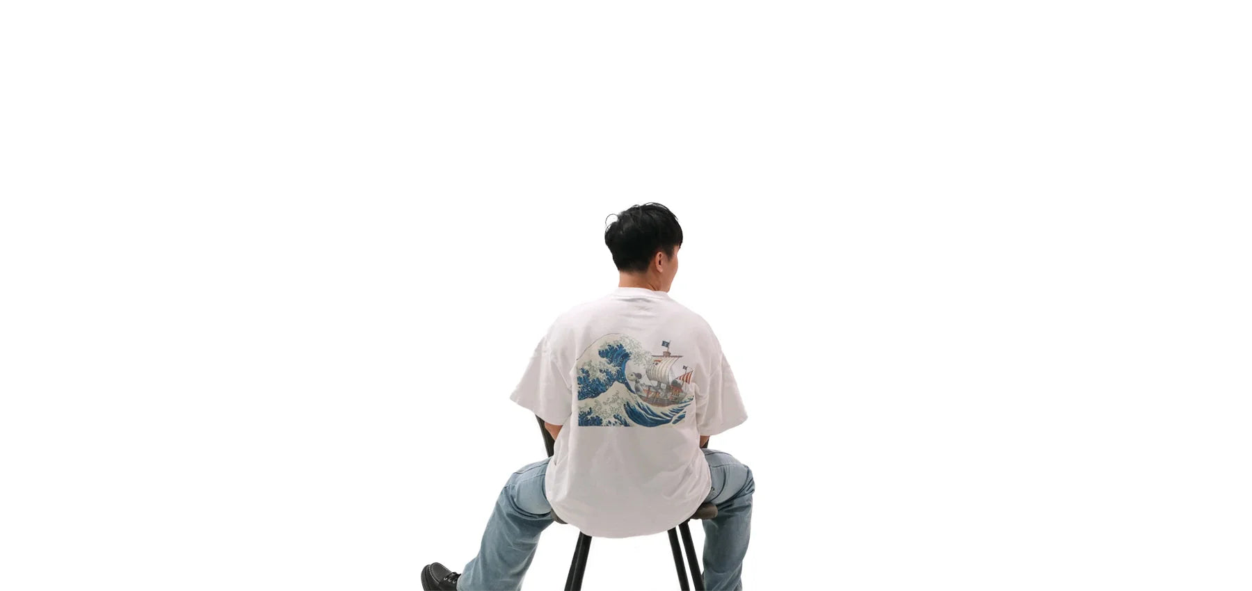 Back view of oversized white NO1 Apparels anime t-shirt, subtle anime clothing inspired by One Piece Going Merry ship with Kanagawa Great Wave motif, worn by a seated male model.