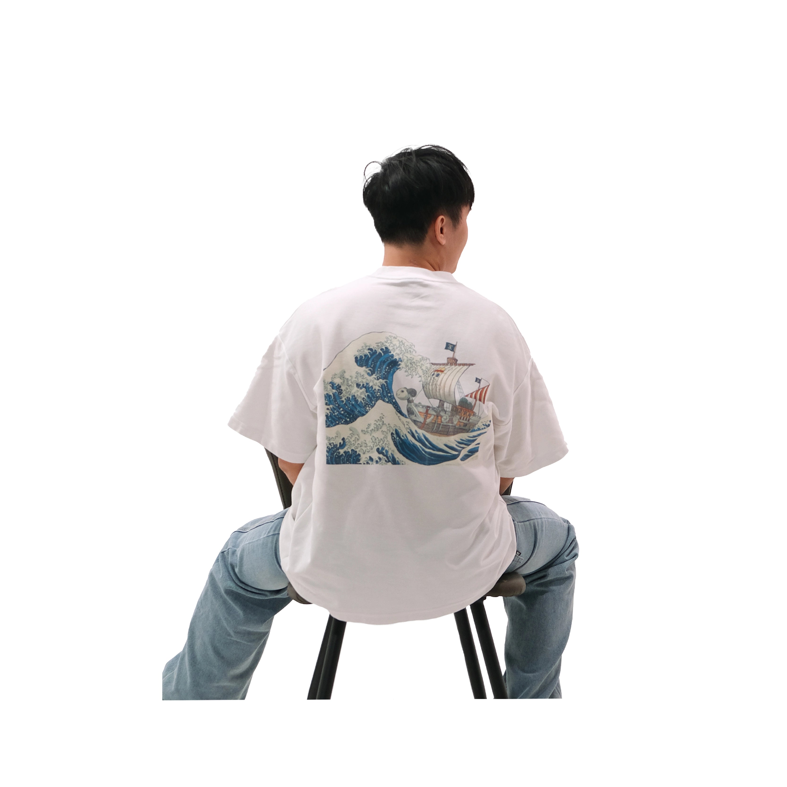 Back view of oversized white NO1 Apparels anime t-shirt, subtle anime streetwear inspired by One Piece Going Merry design featuring ship on The Great Wave, worn by male model sitting on chair.