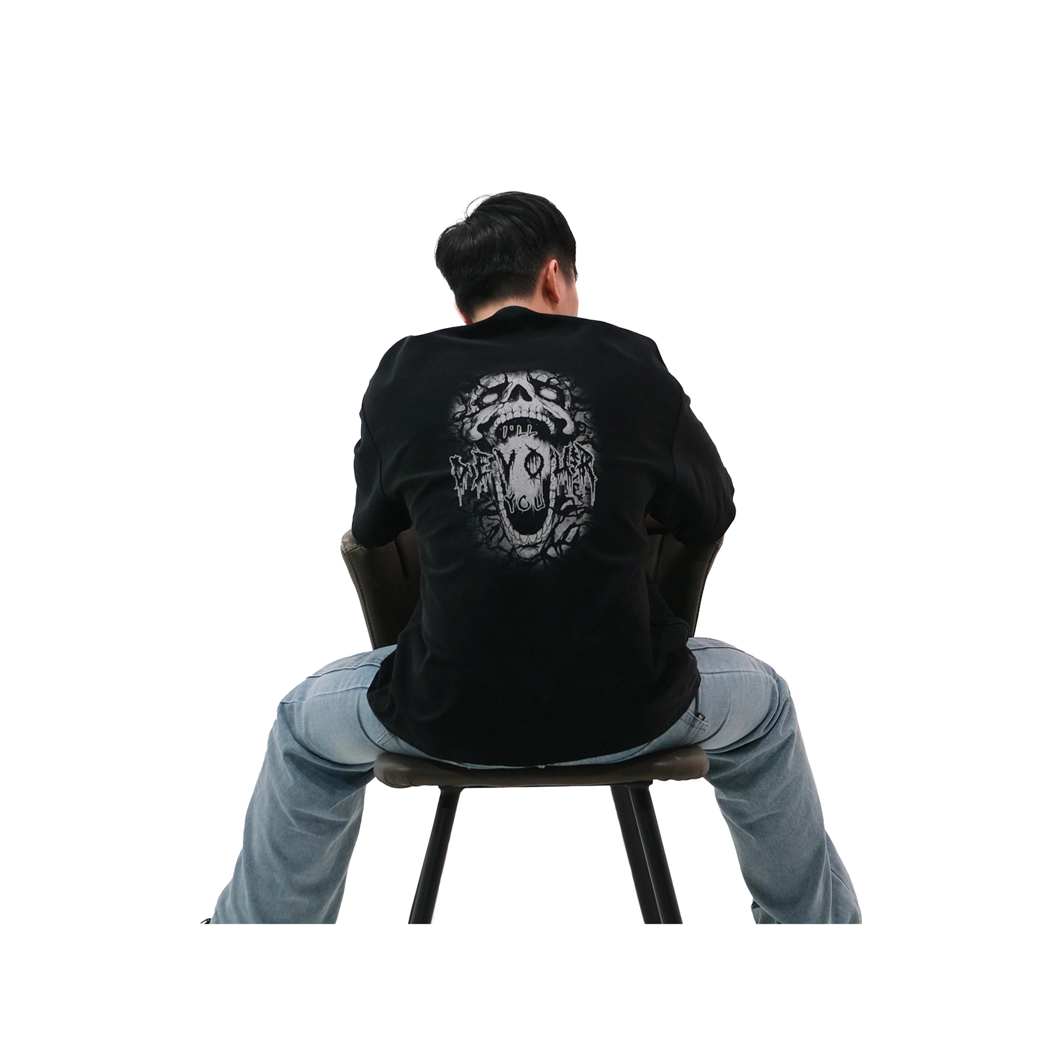 Back view of oversized black NO1 Apparels anime t-shirt, subtle anime streetwear inspired by Blue Lock, with Devour design featuring skull and shadowy motif, worn by male model sitting on chair.